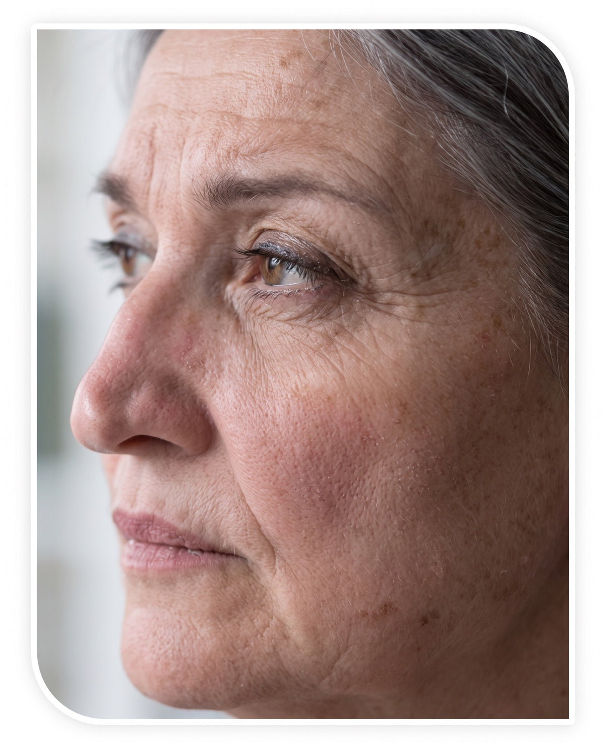 A woman around 48 at her bathroom mirror in morning light, one hand lightly lifting the skin at her jaw, the way every w